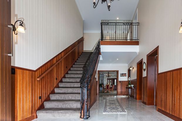 A grand residential entry hall featuring a carpeted staircase with dark ornate metal balustrades, rich timber wall panelling and wainscoting, pale vertical wall panelling above, polished light stone-look flooring, and a small upper-level landing. Decorative wall sconces and a chandelier add traditional character, with a dining area visible at the far end.