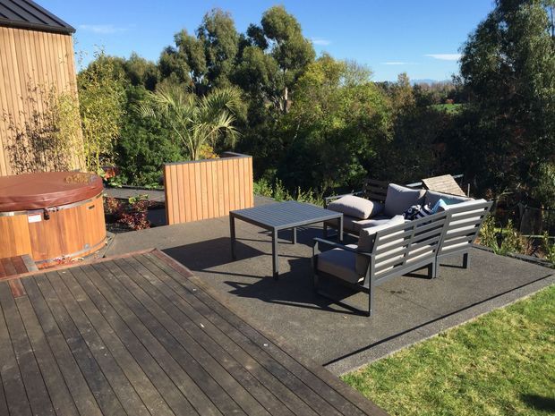 An elevated outdoor terrace with dark timber decking and a paved seating area, furnished with a grey outdoor lounge set and coffee table. A round timber-clad spa sits to the left, with a timber privacy screen and lush planting surrounding the deck. The scene is sunny and leafy, with distant hills visible beyond the garden.