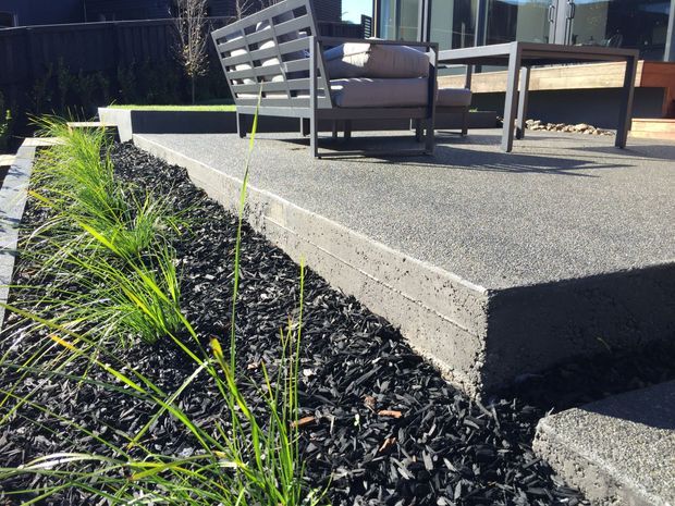 A contemporary landscaped outdoor terrace with a raised textured concrete deck, dark mulch garden bed, ornamental grasses, and modern outdoor lounge furniture beside large glazed doors. The space uses strong geometric lines and dark neutral finishes, with a small gravel feature area integrated near the seating.