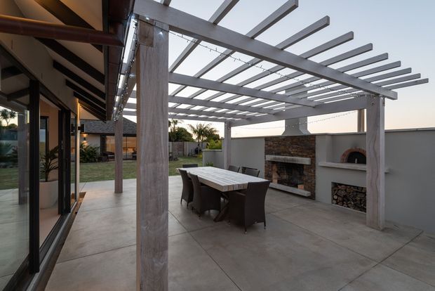 A contemporary covered outdoor entertaining area beside a glazed interior space. The patio features a large concrete-paved floor, timber or timber-look posts and beams forming an open pergola, a dining table with dark outdoor chairs, and a rendered masonry wall incorporating a built-in fireplace, wood storage, and pizza oven. The space opens onto a landscaped garden and lawn.