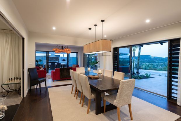 Open-plan dining and living area with a large dark timber dining table, upholstered dining chairs, pendant lighting, dark timber flooring and a light neutral rug. Floor-to-ceiling glazing and wide sliding doors open to an outdoor terrace with a scenic suburban or coastal landscape beyond. A living room with red seating is visible through the opening, and a glass-fronted display area with a chess set appears at left.