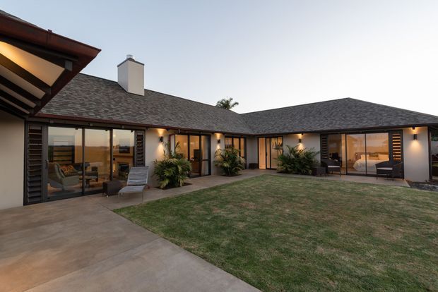 Single-storey courtyard-style residence viewed from the outdoor lawn and concrete path, with a low-pitched dark shingle roof, pale rendered walls, extensive glazed doors and windows, timber or dark slatted privacy screens, and warm interior lighting. Several furnished rooms are visible through the glazing, while the main subject is the building exterior and enclosed courtyard.