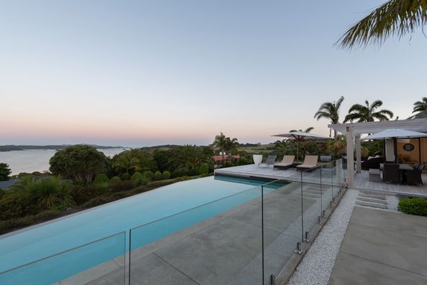 A contemporary outdoor pool terrace overlooking a coastal or waterfront landscape at dusk. The long turquoise pool is bordered by concrete paving and clear glass balustrades, with lounge chairs, a covered outdoor entertaining area, palms, and sculpted landscaping. The setting has an open, resort-like atmosphere with expansive views.