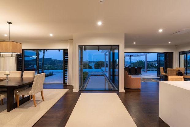 Open-plan contemporary interior with a dining area, kitchen island, and adjoining living space arranged around extensive floor-to-ceiling glazing. Dark timber flooring contrasts with cream rugs, light walls, and warm concealed and pendant lighting. The glazing opens toward an outdoor terrace and landscaped coastal or suburban view.