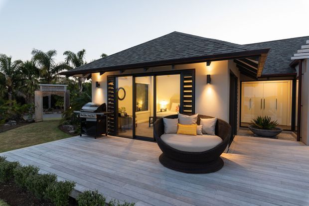 Twilight exterior view of a contemporary single-storey dwelling with a broad dark hipped roof, pale rendered walls, black-framed glazed doors and windows, and a generous timber deck. The deck includes a built-in outdoor barbecue, a large rounded lounge chair, and a low planter. Warm interior and exterior lighting creates an inviting connection between the outdoor entertaining area and the rooms behind the glazing.
