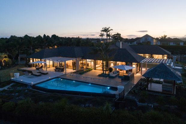 Twilight aerial view of a substantial single-storey residence with dark pitched roofs, extensive illuminated glazing, a large swimming pool, paved terraces, outdoor seating, and lush landscaped gardens. A covered outdoor pavilion and pergola-like structures extend around the pool.