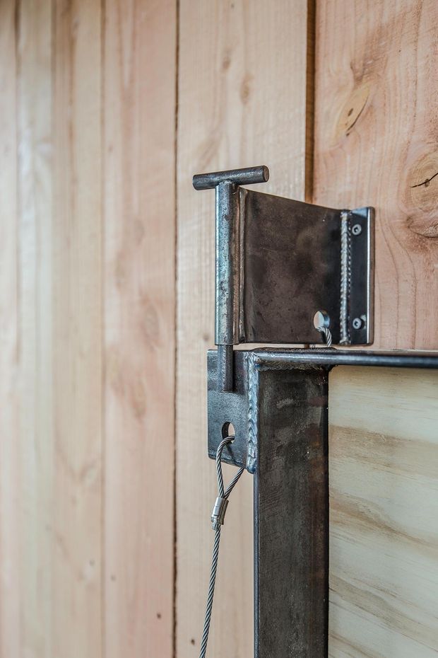 Close-up detail of a rustic sliding barn-style door mechanism, showing a dark raw steel hinge and bracket with a cable latch against vertical natural timber boards. The image focuses on exposed hardware, welds, wood grain, and textured industrial detailing.