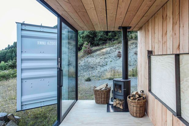 A compact timber-clad covered outdoor living area with full-height black-framed glazing, a timber-lined ceiling, pale timber flooring, and a freestanding wood-burning stove with firewood baskets. A large open shipping-container-style door is visible on the left, while the space opens toward a wooded, sloping landscape.