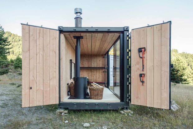A compact converted shipping container or modular cabin set in a natural, wooded landscape. The wide double doors are open to reveal a timber-lined interior with a wood-burning stove and chimney, a wicker basket, shelving, and a glazed side wall or door. Dark steel framing and hardware contrast with the pale timber cladding and interior lining.