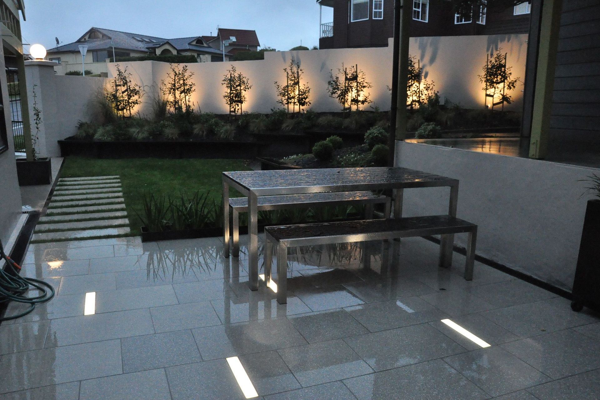 View of garden at night features bright rectangular in ground slot lights and trees uplit along fence line.