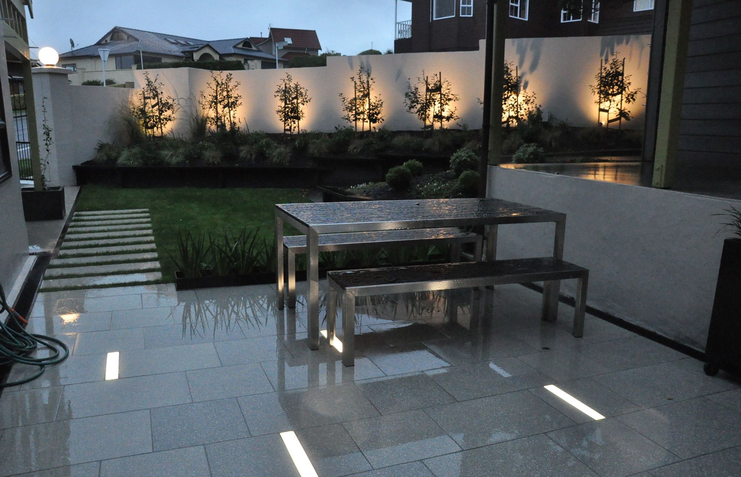 View of garden at night features bright rectangular in ground slot lights and trees uplit along fence line.