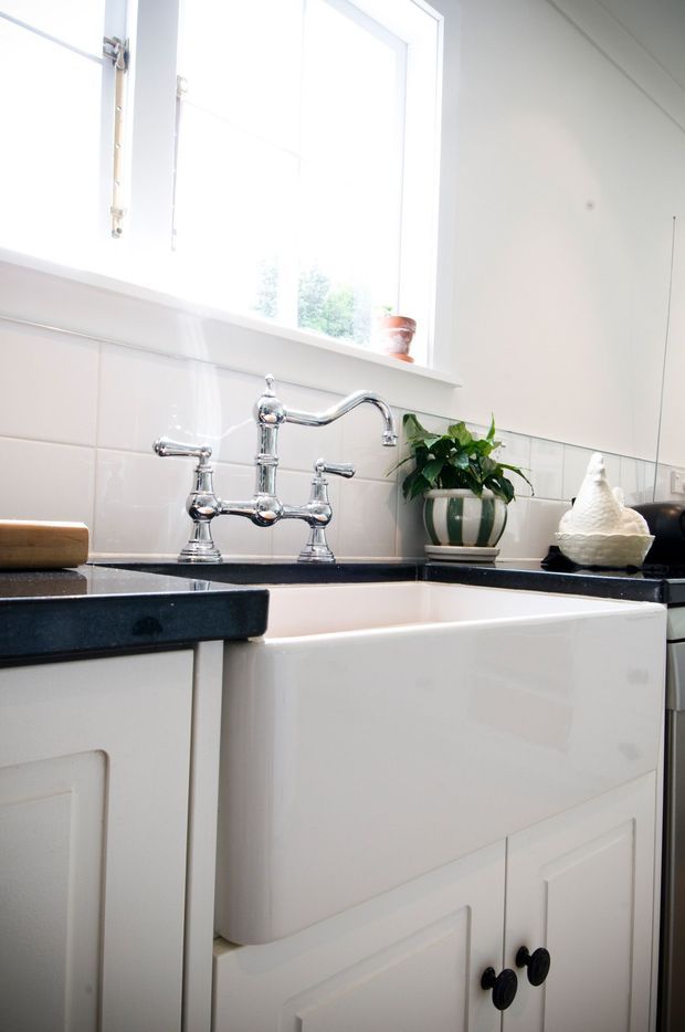 Close-up view of a classic white kitchen sink beneath a bright window. The kitchen features glossy white panelled cabinetry, a dark polished stone benchtop, white ceramic tiled splashback, and a traditional chrome bridge tap with separate handles. Small decorative items and a potted plant sit on the benchtop and windowsill.