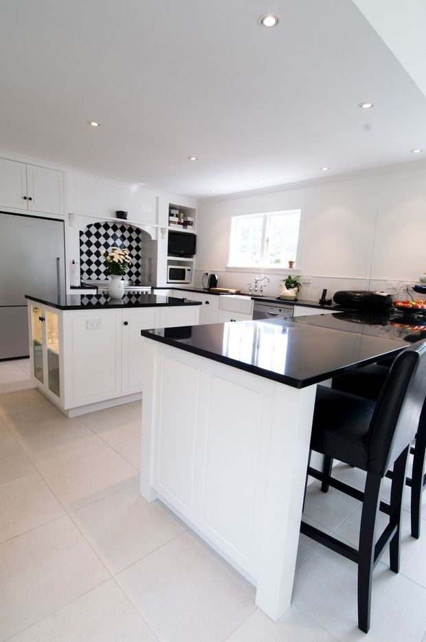 Bright, spacious white kitchen with extensive shaker-style cabinetry, black polished benchtops, a large central island with bar seating, integrated stainless-steel refrigerator, built-in microwave and oven, farmhouse-style sink, and a striking black-and-white checkerboard splashback. Recessed ceiling lights and a large window provide ample illumination.