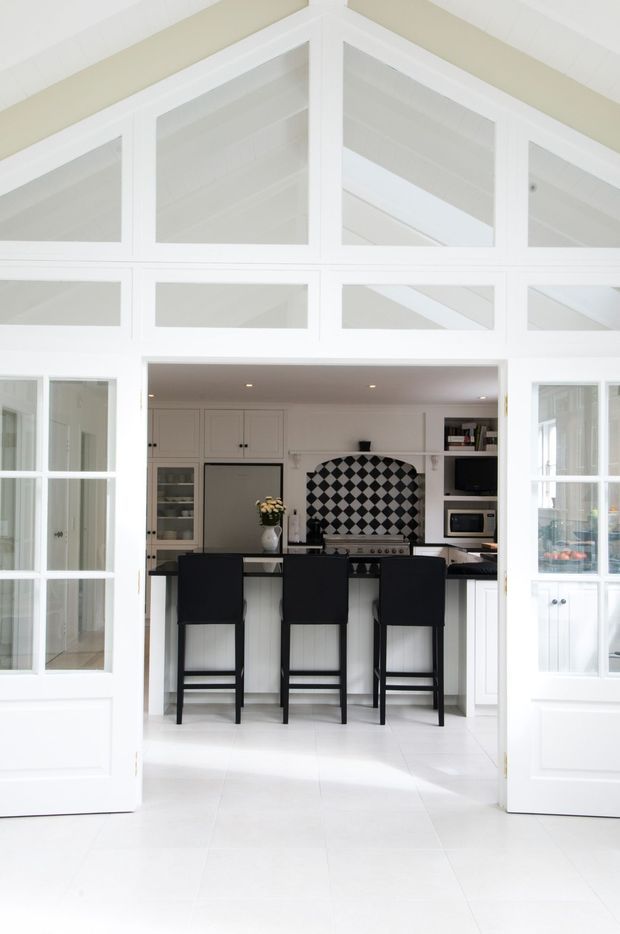 Bright white kitchen viewed through wide glazed double doors, featuring a central island with three black bar stools, white cabinetry, dark benchtops, a geometric black-and-white tiled splashback, and integrated appliances. The room has abundant white framing, glazed partitions, and a high glazed transom with visible ceiling structure, creating an airy, refined contrast between white architectural elements and black accents.