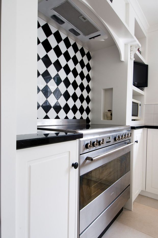 Close-up view of a refined traditional kitchen focused on a large stainless-steel range cooker. The kitchen features white framed cabinetry with black knobs, black stone-look benchtops, and a bold black-and-white diamond checkerboard tile splashback. A sculpted white arch with decorative corbel detailing frames the cooking zone, while a microwave and wall-mounted television are visible in the surrounding joinery.