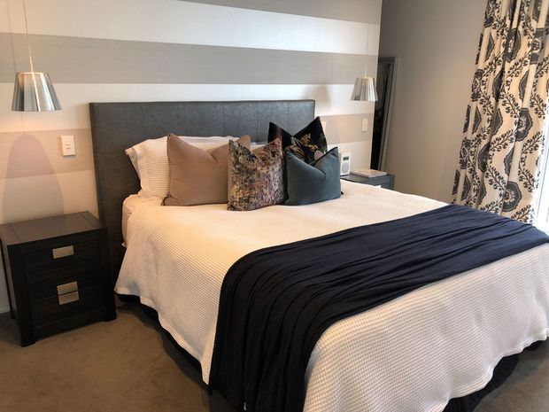 A well-appointed bedroom featuring a large upholstered dark-grey headboard, white textured bedding, layered neutral and patterned cushions, and a dark navy throw. Matching bedside tables with metallic drawer pulls flank the bed, while wall-mounted pendant lights provide symmetrical illumination. The room has soft grey walls, beige carpet, and patterned floor-length curtains.