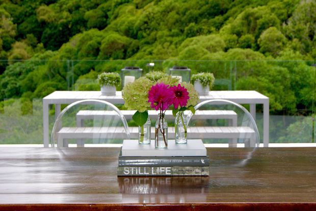 A polished dark timber dining table is arranged with stacked books and a vase of bright pink and green flowers. Behind it, a white outdoor-style dining table and transparent curved chairs sit beside full-height glazing, overlooking dense green vegetation. The scene is bright, refined, and strongly connected to the surrounding landscape.