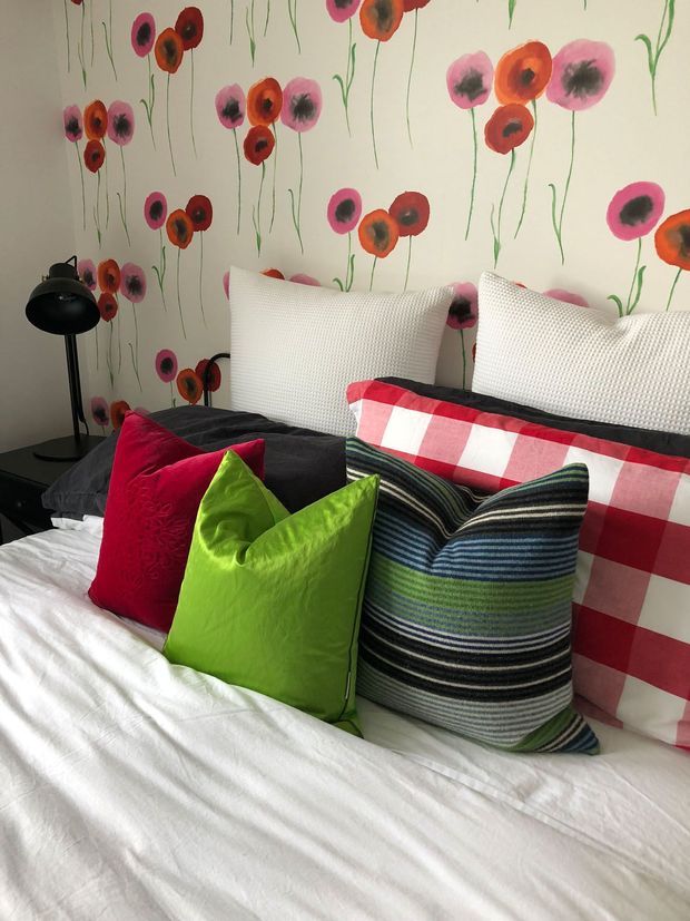 A bright, playful bedroom featuring a white bed layered with dark bedding and colourful decorative cushions in red, lime green, and striped blue-green tones. The wall behind the bed has a hand-painted poppy floral mural, while a black floor lamp and dark bedside furniture sit to the left.