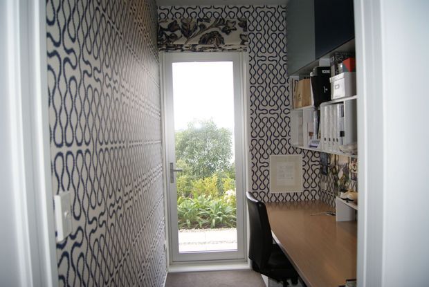 A narrow home office with a built-in timber desk and black office chair, wall-mounted open shelving and storage, and highly distinctive white wallpaper featuring repeating navy-blue loop patterns. A tall glazed door or window at the end provides views of greenery and brings in natural light. The space is compact, functional, and visually bold.