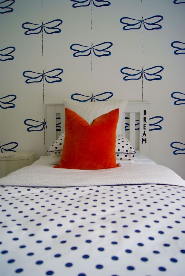 A playful bedroom centered on a neatly made single bed. White bedding with navy-blue polka dots and a bright orange accent cushion contrasts with white furniture and wallpaper featuring repeated blue dragonfly motifs. The white bedhead includes the word “DREAM” cut into or printed on its vertical slats, creating a cheerful child-friendly look.