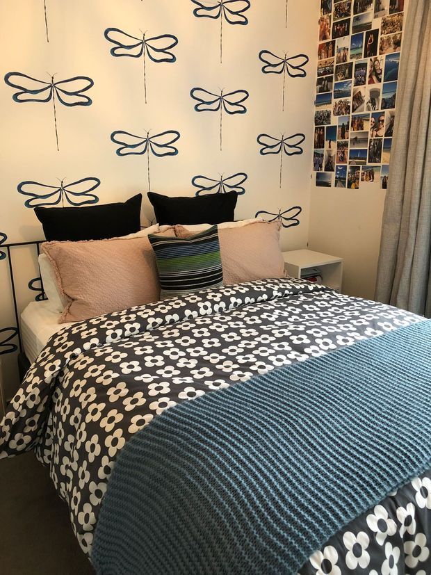 A compact, playful bedroom centered on a double bed with layered patterned bedding, pale pink pillows, black cushions, and a striped accent cushion. The cream walls feature repeated hand-drawn navy butterfly motifs, while a collage of personal photographs decorates the upper right wall. A small white bedside table and full-height curtains complete the room.