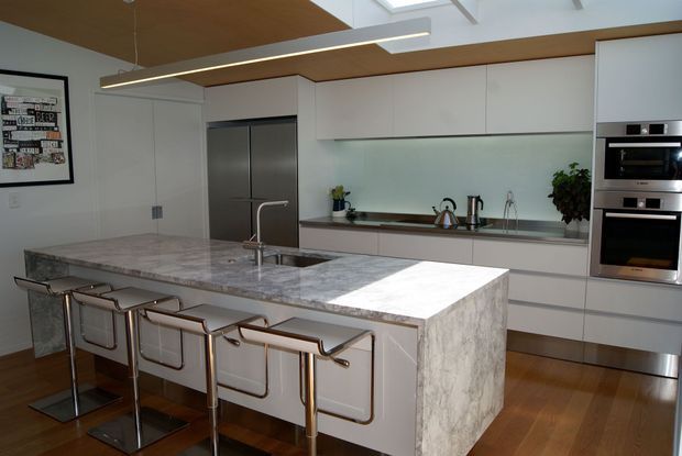 A bright contemporary kitchen featuring a large marble-look waterfall island with integrated sink and four bar stools, flat-panel white cabinetry, stainless-steel appliances, a pale glass backsplash, timber flooring, and a timber-lined ceiling with skylights and linear pendant lighting.