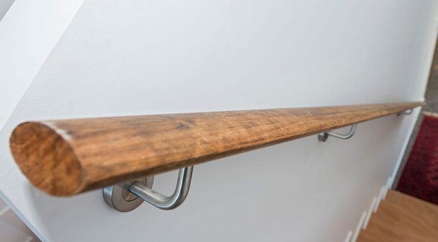 Close-up detail of a wall-mounted timber handrail with a rounded end, supported by curved brushed-metal brackets against a white wall. A timber stair or landing edge and a red carpeted area are partially visible at the right.