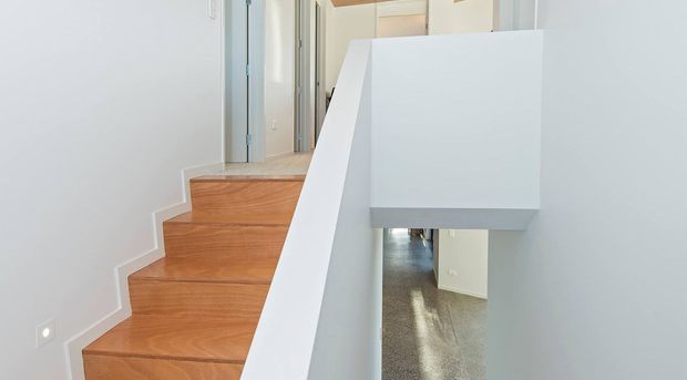 Bright contemporary stairway with warm timber treads, crisp white walls and balustrade, and a view down to a polished grey floor below. The minimalist detailing and clean geometric form create an airy, modern interior.