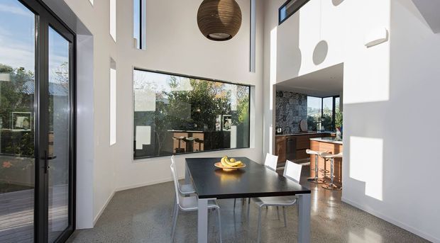 Bright contemporary open-plan dining area with a black rectangular dining table and white chairs, polished concrete or terrazzo-style flooring, white walls, large black-framed windows and doors, and a dramatic double-height ceiling. A compact kitchen is visible beyond the dining space, with strong natural light and a sculptural timber pendant light.