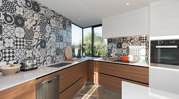 Bright contemporary kitchen with an L-shaped layout, white stone-look benchtops, warm timber cabinetry, integrated stainless-steel appliances, and a striking monochrome patterned tile splashback extending across the main wall and cooktop area. Large corner glazing brings in natural light and views of greenery.