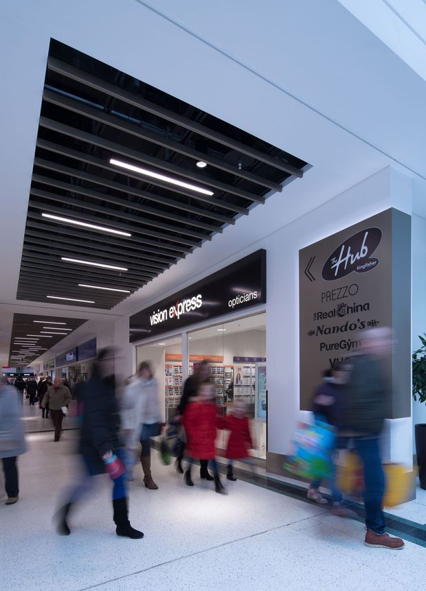 Interior view of a contemporary shopping centre concourse with blurred pedestrians, retail storefronts including Vision Express opticians, a large directory sign, polished light terrazzo flooring, and a striking suspended black slatted ceiling with integrated linear lighting.