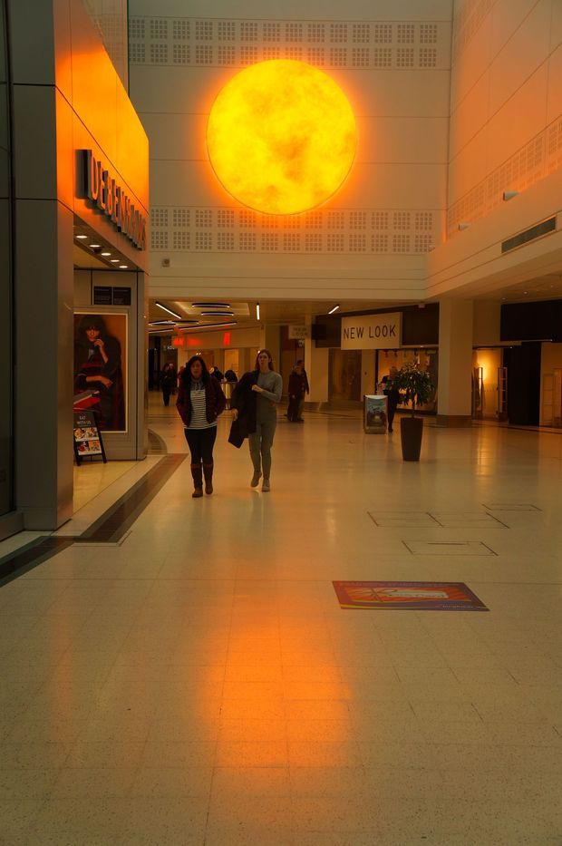 Interior view of a spacious commercial shopping-centre concourse with polished light flooring, retail storefronts, recessed ceiling lighting, perforated wall panels, signage, indoor planting and people walking through the space. A large glowing circular orange-yellow feature light or illuminated artwork dominates the upper wall and casts a warm reflection onto the floor.