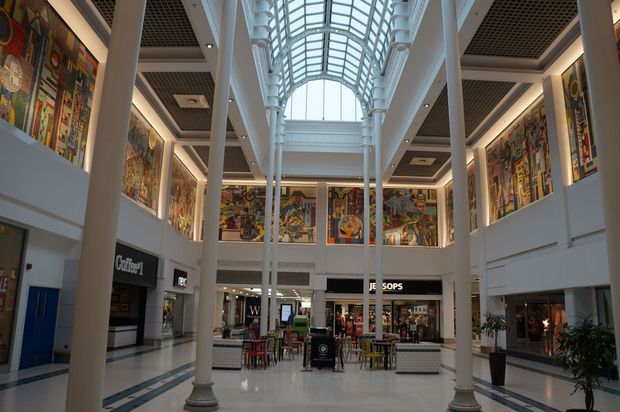 Interior view of a multi-level shopping mall or commercial atrium, featuring tall classical-style columns, a large barrel-vaulted glass skylight, patterned coffered ceilings, polished light flooring, retail storefronts, and colourful large-scale artwork displayed along the upper walls.