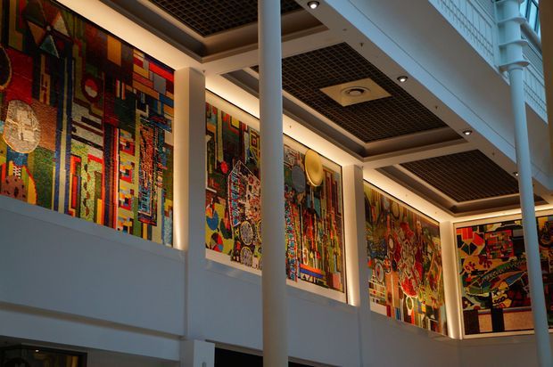 Interior view of a spacious multi-level commercial or public building atrium, featuring tall white structural columns, illuminated abstract multicoloured mural panels lining the upper walls, and a dark recessed grid ceiling with integrated lighting and ventilation. The space has a contemporary institutional or civic character.