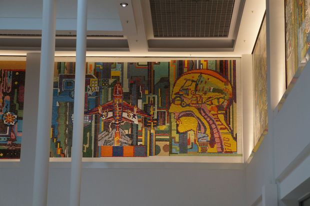 Interior view of a contemporary public or commercial space featuring large colourful geometric mosaic artworks mounted on the walls, visible across an open void or stairwell. White structural columns, clean white walls, recessed ceiling lighting, and a dark gridded ceiling panel frame the artwork.