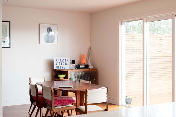 A warm, light-filled dining area featuring a mid-century timber dining table and upholstered dining chairs, with a vintage timber sideboard, framed artwork, and large glazed doors opening to an exterior courtyard or deck.