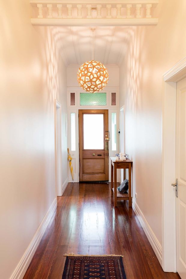 A bright, narrow residential entry hallway with polished dark timber flooring, pale walls, white decorative trim, and a high ceiling. A timber front door with sidelights and coloured leadlight glazing is positioned at the far end. A spherical pendant light casts patterned shadows across the walls, while a slim console table, umbrella stand, and runner rug furnish the corridor. Decorative ceiling cornice and architraves add traditional character.