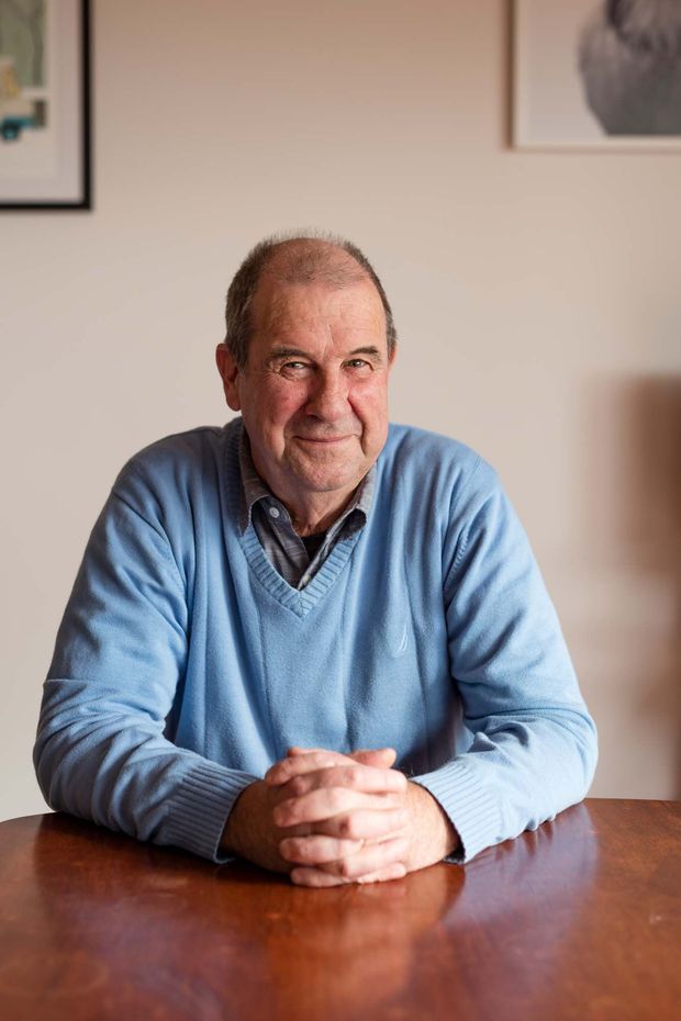 Portrait of an older man seated at a polished wooden table in a softly lit, neutral-toned interior. Framed artwork is visible on the wall behind him, creating a warm and informal home setting.