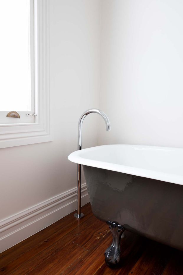 Close-up view of a classic freestanding clawfoot bathtub in a bright bathroom, with a polished chrome floor-mounted tub filler, white decorative wall panelling and trim, a white-framed window, and warm dark timber flooring.