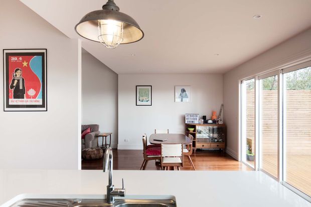 Open-plan interior viewed across a white kitchen island toward a compact dining area and adjoining living space. The room has warm timber flooring, pale walls, large sliding glass doors opening to a timber deck, framed artwork, a timber dining table and chairs, and a dark metal pendant light over the kitchen. The palette is bright and neutral with warm wood accents.