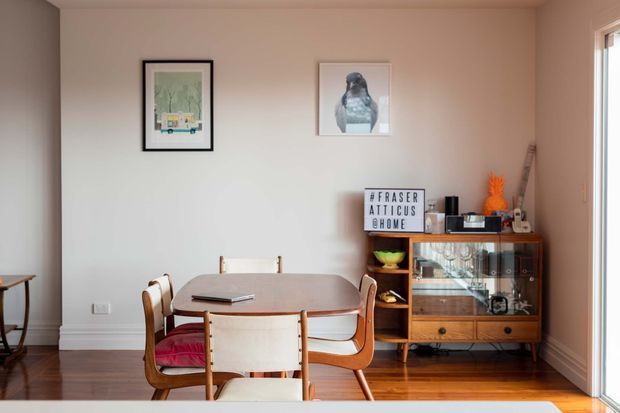 A warm, mid-century-inspired dining room with a rounded rectangular wooden dining table and matching timber chairs upholstered in red. Framed artwork hangs on a pale wall, while a vintage timber and glass display cabinet holds decorative objects. Timber flooring, soft natural light, and restrained decor create an inviting retro atmosphere.