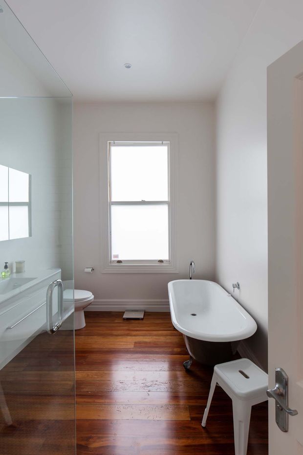 Bright, narrow bathroom with a freestanding white bathtub, white toilet, glass shower enclosure, wall-mounted vanity, and a small stool. The room features warm polished timber flooring, pale neutral walls, white trim, and a central window providing strong natural light.