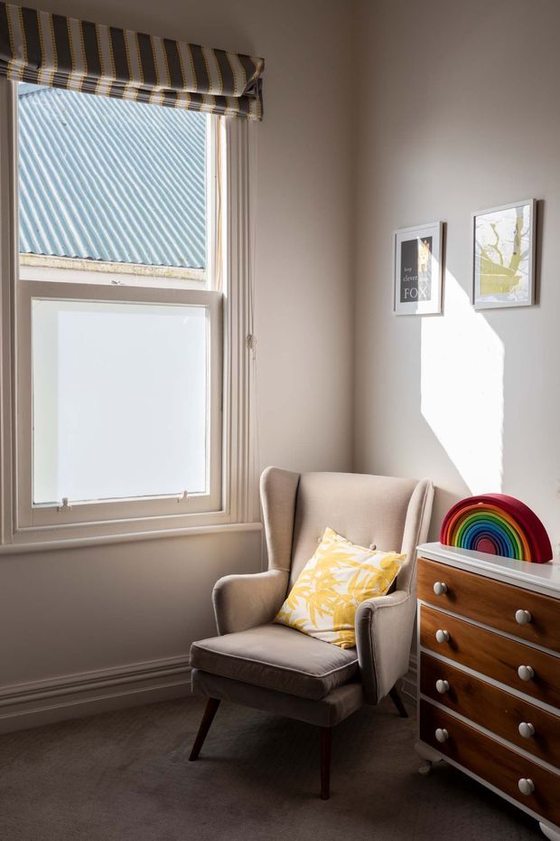 A small, warmly lit sitting corner with a beige upholstered wingback armchair, patterned yellow cushion, and a mid-century-style wooden chest of drawers topped with a colourful rainbow ornament. A tall sash-style window with a striped Roman blind occupies the left side, while framed prints hang on the light neutral wall. Carpeted flooring and traditional skirting boards complete the intimate interior.