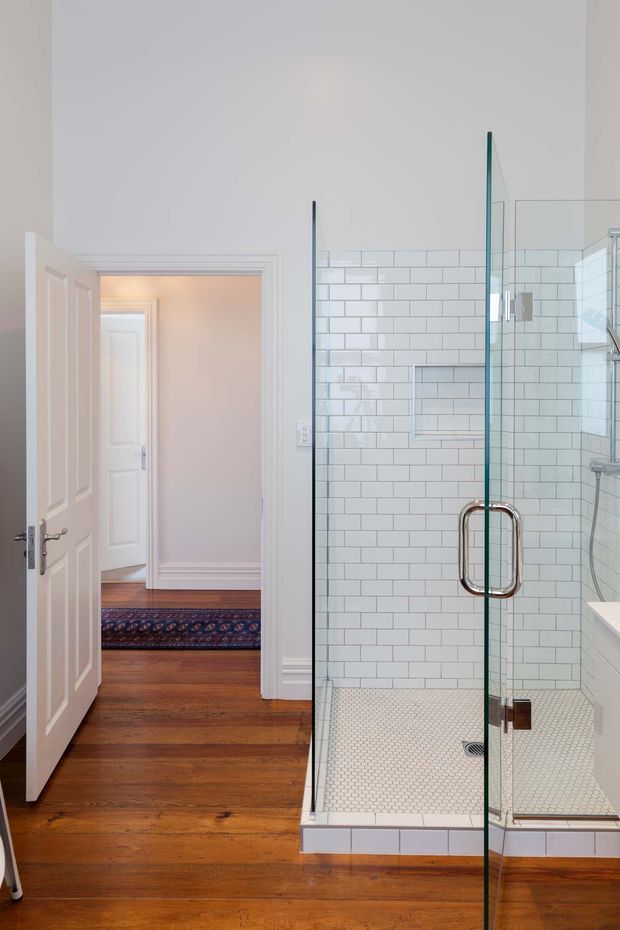 Bright traditional bathroom with a large frameless glass walk-in shower, white subway-tiled walls, small white mosaic shower floor, chrome and polished metal hardware, and white trim. The bathroom opens toward a hallway with warm timber flooring and a patterned runner visible beyond the open door.