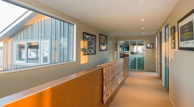 A long, bright residential hallway or landing with warm neutral walls, carpeted flooring, recessed ceiling lights, framed artwork, and a timber half-height balustrade overlooking the level below. Large windows on the left provide views of a modern glazed and vertically clad exterior structure, while several doorways and a glazed door lead toward the far end.