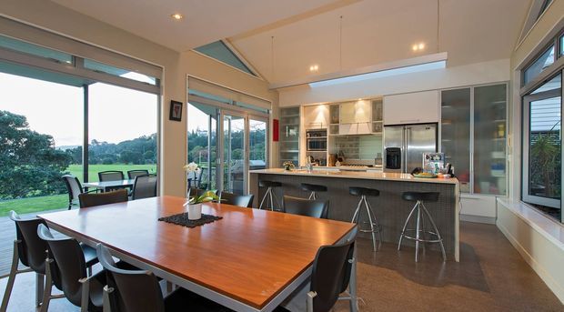 Bright open-plan kitchen and dining area with a large timber dining table, contemporary dark seating, a central island with bar stools, integrated cabinetry and stainless-steel appliances. Extensive windows and glazed doors provide views of landscaped surroundings, while a high angled ceiling and clerestory-style glazing add openness and daylight.