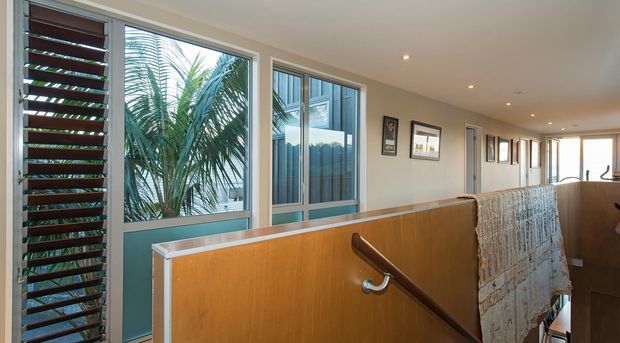 Bright modern interior hallway or stair landing with warm timber half-height balustrade panels, a dark timber handrail, large aluminium-framed windows and a louvred external screen. Framed artwork lines the cream-coloured corridor walls, with tropical planting visible through the glazing.