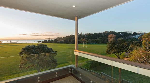 A covered balcony or terrace overlooking expansive sports fields and a calm waterfront at sunset. The space features clear glass balustrades, a timber-toned deck, slim structural posts, and a broad flat roof canopy, creating an elevated outdoor viewing area.
