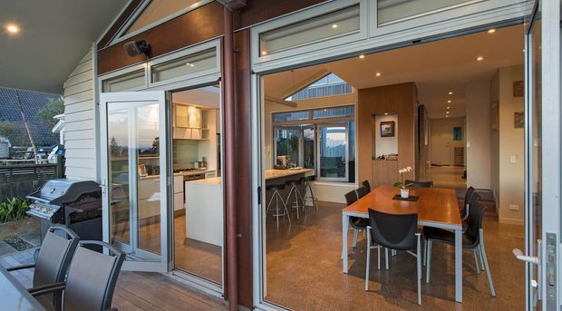View from a covered outdoor deck through wide-open glazed doors into a contemporary open-plan kitchen and dining area. The interior features warm timber wall and ceiling surfaces, polished concrete flooring, a timber dining table with modern chairs, and a kitchen island with stools. A barbecue and outdoor seating are visible on the deck, while large glazed openings connect the indoor and outdoor spaces.