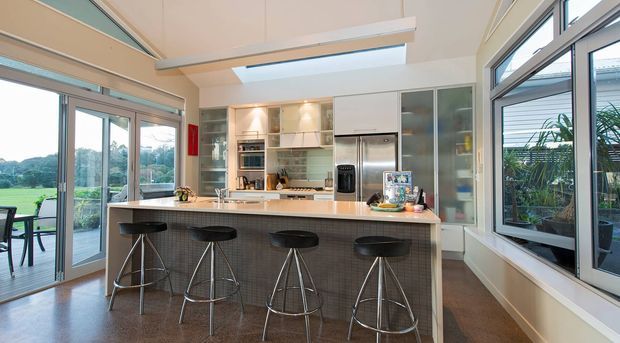Bright, spacious contemporary kitchen with a large island and four bar stools, extensive glazing, sliding doors to an outdoor area, skylight, stainless-steel appliances, and pale built-in cabinetry. The room has a light, airy feel with views to greenery outside.